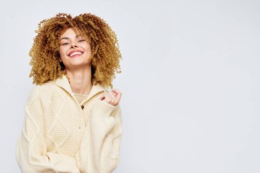 Şehvetli kıvırcık saçlı neşeli bir kadın, yumuşak krem süveter giyiyor, sade bir arka plana karşı güvenle gülümsüyor, neşe, rahatlık ve günlük yaşam tarzı fotoğrafçılığını temsil ediyor.