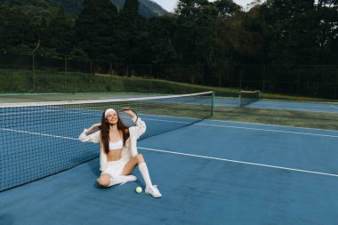 Atletik giyinen neşeli genç kadın, neşeli ifadesiyle enerjik ve sağlıklı bir yaşam tarzını yansıtan tenis kortunda poz veriyor.