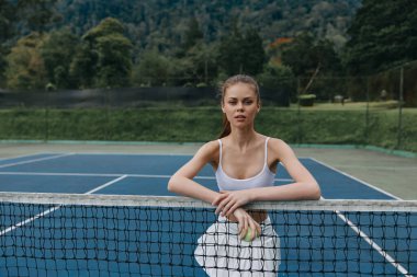 Spor elbiseli kararlı bir genç kadın tenis sahasının yanında duruyor, sakin bir tenis kortunda bir top tutuyor yemyeşil arka plan coşkulu bir atletizm ve özgüven sahnesini geliştiriyor.