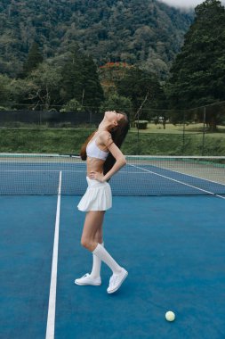 Şık beyaz tenis kıyafeti giymiş kendine güvenen genç bir kadın mavi bir kortta duruyor, yemyeşil dağların arasında kararlılık ve neşe ifade ediyor, spor ve spor temaları için mükemmel.