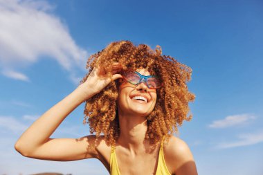 Güzel bukleli neşeli bir genç kadın, sarı bir bluz giymiş, açık havada güneşli bir günün tadını çıkarıyor.