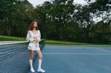 Rahat genç bayan tenis oyuncusu şık spor kıyafetleriyle sahada poz veriyor, tenisin doğasını ve neşesini kucaklıyor, sakin ve odaklanmış bir ifadeyi sergiliyor