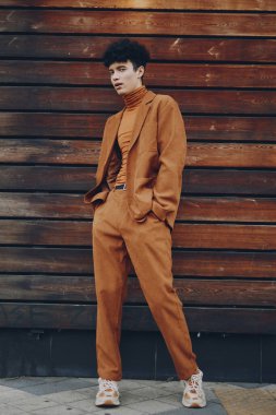 Turuncu kahverengi bir takım elbise ve balıkçı yaka giyen modaya uygun bir erkek model, kırsal bir ahşap zemine karşı poz veriyor.