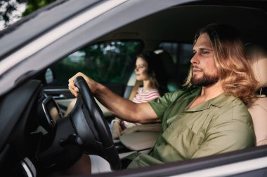 Uzun saçlı genç bir adam kendinden emin bir şekilde araba kullanırken yanında oturan bir kadın yolculuğun tadını çıkarıyor.