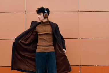 Turuncu bir duvarın karşısında duran, kulaklıklı, şık bir genç adam. Kahverengi bir kazak, mavi kot pantolon ve kahverengi ayakkabılar giyiyor..