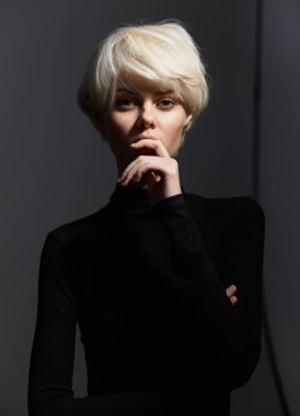 Modern saç kesimi olan şık bir genç kadın. Forma uygun siyah bir elbise içinde dalgın bir ifade sergiliyor. Işığın ve gölgenin kontrastı onun özelliklerini vurgular.