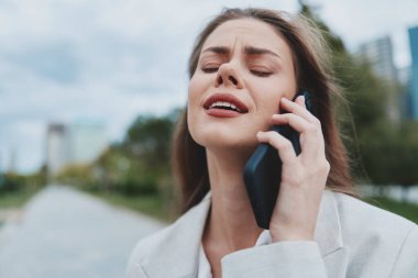İş kıyafetleri içinde, açık mavi gökyüzü olan bir şehir parkında telefonla konuşurken duygusal hisseden, modern profesyonel hayatın zorluklarını gözler önüne seren etkileyici genç bir kadın..