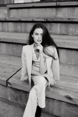 Profesyonel bir iş kadını basamaklarda otururken, çağdaş bir kıyafetle güven yayıyor. Bu siyah beyaz görüntü, modern kurumsal kültürün ve güçlenmenin özünü yansıtıyor..