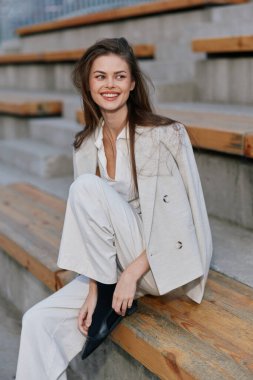 Uzun saçlı, kendine güvenen bir iş kadını ahşap basamaklarda otururken gülümser, şık beyaz bir kıyafet giyer, modern profesyonelliği çağdaş bir ortamda somutlaştırır..
