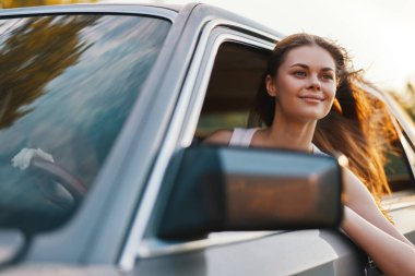Neşeli genç bir kadın, yolculukta özgürlüğünü yaşıyor. Arabasının camından eğilmiş, saçları uçuşuyor. Macera ve mutluluğun özünü yakalıyor..
