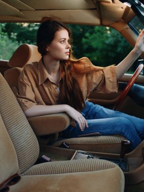 Büyük bej gömlekli ve kot pantolonlu genç bir kadın antika bir arabada oturmuş, pencereden dışarı dikkatlice bakıyor, yemyeşil bir arka plan, nostalji ve özgürlüğü çağrıştırıyor..