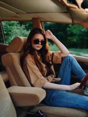 Uzun saçlı, güneş gözlüğü takan ve rahat bir gömlek giyen güzel genç bir kadın, klasik bir arabada rahatça oturuyor, özgürlük ve stil duygusu sergiliyor..