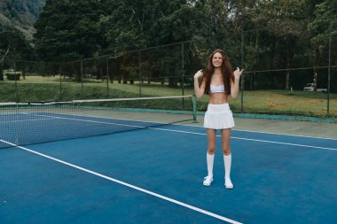 Beyaz spor giyimli enerjik bayan sporcu yeşil ağaçlarla çevrili mavi tenis kortunda neşe saçıyor. Yaz sporları ve spor için ideal.