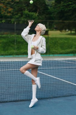 Beyaz tenis kıyafetli şık bir genç manken, sahada oyun oynamaktan zevk alıyor. Canlı yeşil bir arka plana karşı güven ve mutluluk yayıyor.