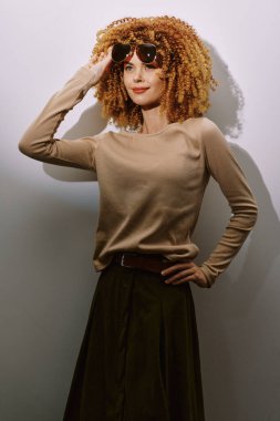 Kıvırcık saçlı, güneş gözlüğü takan şık bir kadın tarafsız bir arkaplanda poz verir. Kıyafeti, açık renk bir kazağı ve koyu renk bir eteği vardır. Taze ve şık bir tarzı yakalar.