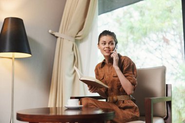 Kahverengi tulum giymiş genç bir kadın, modern bir iç mekanda huzur dolu bir anın tadını çıkarırken telefonda konuşuyor ve bir kitap okuyor, dinginlik ve tarz yayıyor.