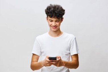 Gülümseyen genç adam akıllı telefonuyla meşgul, basit beyaz bir tişört giyiyor açık gri arka plan modern bir ortam ekliyor, günlük hayatta bağlantı ve teknolojinin temasını vurguluyor.