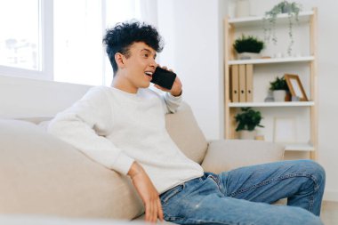 Genç adam parlak bir oturma odasında bej bir kanepede uzanırken telefon konuşmasından zevk alıyor. Alan modern dekorasyon ve bol doğal ışık barındırıyor. Rahat ve davetkar bir atmosfer yaratıyor.