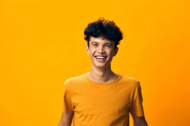 Kıvırcık saçlı neşeli bir genç, parlak sarı bir arkaplanda gülümsüyor, günlük turuncu bir tişört giyiyor, mutluluk ve pozitiflik yayıyor, canlı, güneşli bir atmosferi somutlaştırıyor.