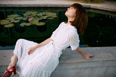 Güneşli bir günde çiçekli nilüferlerle gölün kenarında oturan, beyaz elbiseli sakin bir kadın.