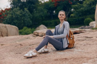 Rahat giyimli genç bir kadın kayalık arazide oturuyor, canlı, yeşil bir zemine hafifçe gülümsüyor. Bu resim rahatlama ve açık hava macerası hissini tasvir ediyor.