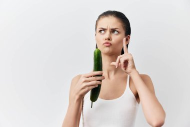 Bir elinde salatalık tutan genç bir kadın parmağını kaldırmış, temiz beyaz bir arkaplana karşı oynayarak neşeli bir yüz ifadesi sergiliyor.