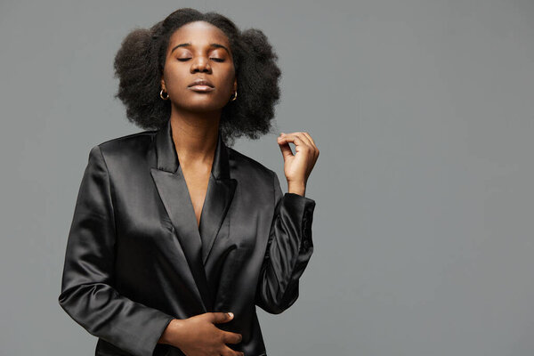 Stylish young black woman in a black blazer, showcasing natural hair and a calm demeanor The minimalistic gray background emphasizes confidence and modern fashion