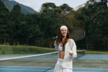 Beyaz spor kıyafetleri giyen neşeli bayan tenisçi, güneşli bir kortta tenis topu tutuyor, coşku ve canlılığı yakalıyor, spor ve spor teması için ideal.