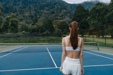 Beyaz atletik kıyafetli kararlı genç bayan tenisçi, mavi bir tenis kortunun üzerinde, yemyeşil dağlardan oluşan bir zemin ile, spor ve zindeliği temsil ediyor.