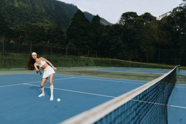 Sportif beyaz elbiseli genç bir bayan tenisçi, coşkuyla dolu, mavi bir tenis kortunda duruyor, yemyeşil dağlık bir arka planda, somut bir atletik enerji.