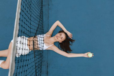 Beyazlar içindeki genç bayan tenis kortu maçında yalan söylüyor, elinde bir tenis topu tutuyor, canlı mavi bir arka planda rahatlık ve güven yayıyor.