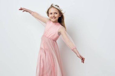 Narin pembe tül bir elbise ve çiçek desenli bir saç bandı takan mutlu bir genç kız yumuşak, nötr bir zemin üzerinde neşeyle gülüyor ve dans ediyor. Çocuk modası ve yaşam tarzı teması için ideal.