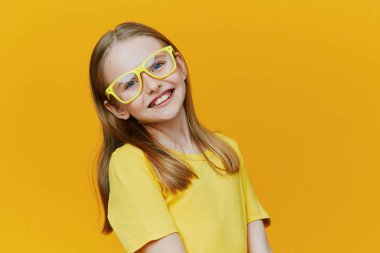 Gözlüklü, sarı tişörtlü neşeli bir genç kız çarpıcı sarı bir arka plana sahip, mutluluk ve kendine güveni somutlaştıran, çocuk modası ve neşeli temalar için ideal.