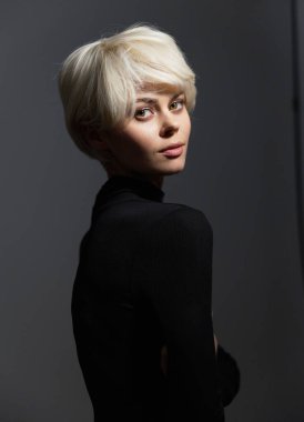 Kendine güvenen, şık, platin sarısı saçlı, siyah elbiseli, omzunun üzerinden bakan genç bir kadın. Dramatik ışıklandırma büyüleyici özelliklerini geliştirir..