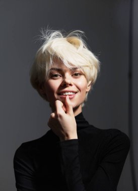 Kısa sarı saçlı, siyah balıkçı yaka giyen, gülümseyen ve şakacı bir şekilde çenesine dokunan kendine güvenen genç bir manken. Minimalist stüdyo ortamında parlak bir ifade.