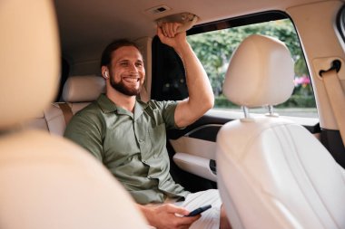 Sıradan giyinmiş bir adam arabada oturuyor, müzik dinlerken gülümsüyor, yumuşak bir iç mekanla çevrili.