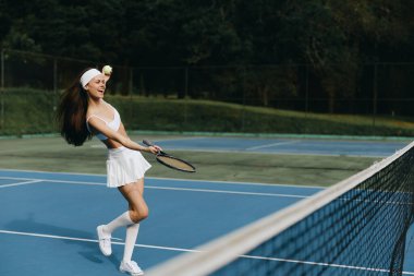 Genç ve neşeli bir bayan tenisçi tenis kortunda topa vuruyor, sportif bir kıyafet giyiyor canlı mavi ve yeşil renkler, yaz sporları için mükemmel bir ortam oluşturuyor.