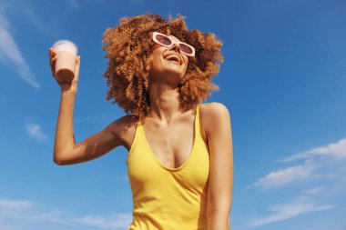 Sarı mayo ve güneş gözlüğü takmış neşeli bir kadın, güneşli mavi bir arka plana gülümseyen bir smoothie tutuyor.