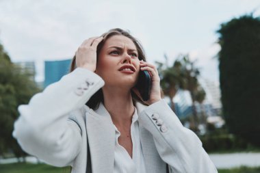 Şık gri ceket giymiş endişeli bir iş kadını şehir dışında bir yerde telefonla iletişim kuruyor. Sahne, modern bir profesyonelin duygusal gerilimini ve stresini yansıtıyor.
