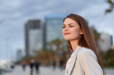 Şık bir bluz giymiş, gün batımında şehir binalarının önünde gülümseyen, profesyonelliği ve iş tutkusunu simgeleyen kendine güvenen bir kadın..