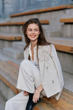 Açık renkli iş kıyafeti giymiş kendine güvenen bir kadın açık havada oturuyor, sıcak bir gülümsemeyle pozitiflik yayıyor, modern bir şehir zeminine karşı kurulmuş..