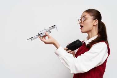 Gözlüklü genç bir kadın bir elinde küçük bir dron, diğer elinde uzaktan kumandayla beyaz bir arka planı olan modern bir ev ortamını gösteriyor.