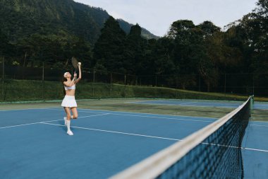 Mavi kortta tenis oynayan neşeli genç bir kadın, spor kıyafetleri içinde, heyecan ve enerji sergiliyor, açık hava sporlarının ve fitnesinin özünü mükemmel bir şekilde yakalıyor.