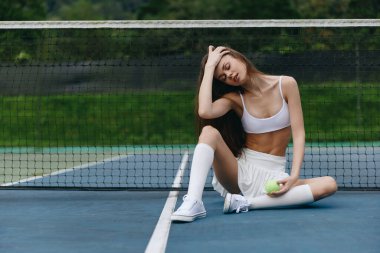 Spor sutyenli ve şortlu genç bir kadın tenis kortunda oturuyor, elinde bir tenis topu, gevşeme ve konsantrasyon sergiliyor.