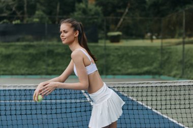 Beyaz tenis kıyafetli formalı genç bir kadın zarif bir şekilde bir baloya hazırlanıyor, canlı bir açık hava kortunda özgüven ve kararlılığı somutlaştırıyor.