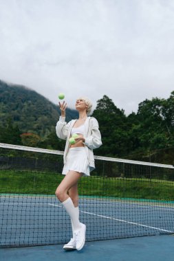 Sahada tenis toplarıyla hokkabazlık yapan enerjik genç bir kadın yeşil zemin ile aktif yaşam tarzını, neşesini ve atletizmini vurguluyor.