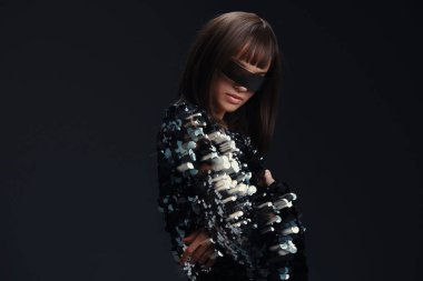 Siyah beyaz elbiseli kadın, gözleri bağlı, kolye ve başörtüsü takıyor, tarafsız bir arkaplanda poz veriyor, gizem, moda ve entrika temalarını çağrıştırıyor.