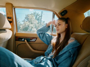 Kot pantolon giymiş düşünceli genç bir kadın klasik bir arabanın içinde oturur ve pencereden dışarı bakar. Parlak gün ışığı onun sakin ifadesini ve şık görünümünü vurguluyor..