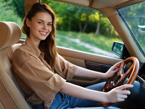 A young woman with long hair joyfully drives a classic car, dressed in a casual beige shirt and blue jeans. The sunny outdoor setting reflects a sense of freedom and adventure, ideal for capturing mo