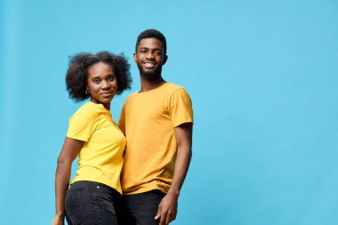 Sarı t-shirtler giyen neşeli bir Afrikalı çift canlı bir mavi zemine karşı birlikte poz vererek çağdaş ve renkli bir atmosferde aşkı ve mutluluğu ifade ediyorlar.